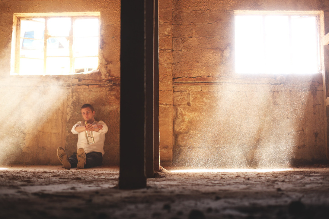 A man sitting on the ground in an empty room, dust visible from the light streaming through the windows